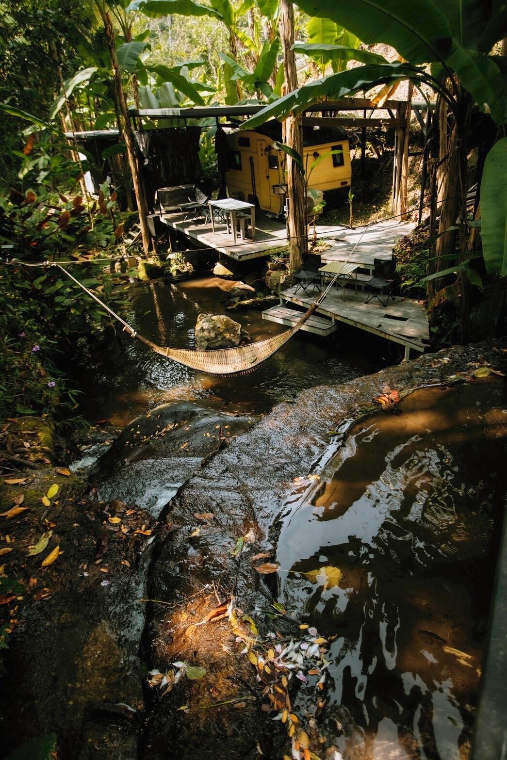 Eine ländliche Außenansicht mit einer Holzbrücke über einem Bach, umgeben von üppiger Vegetation und einem kleinen gelben Gebäude.