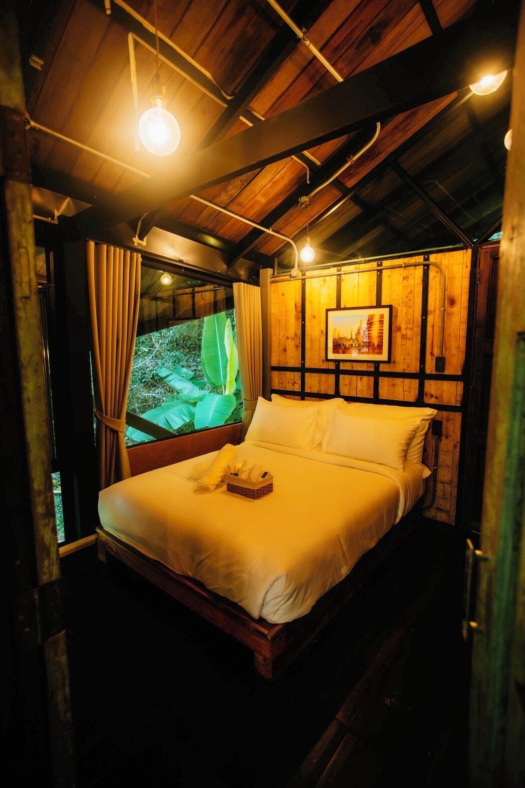 A wooden room with a bed, a framed picture on the wall, and a window with curtains.