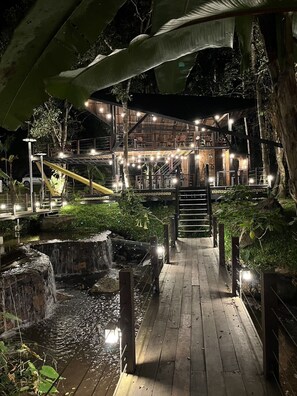 A multi-level wooden structure with outdoor seating, a waterfall, and a wooden pathway leading to it.