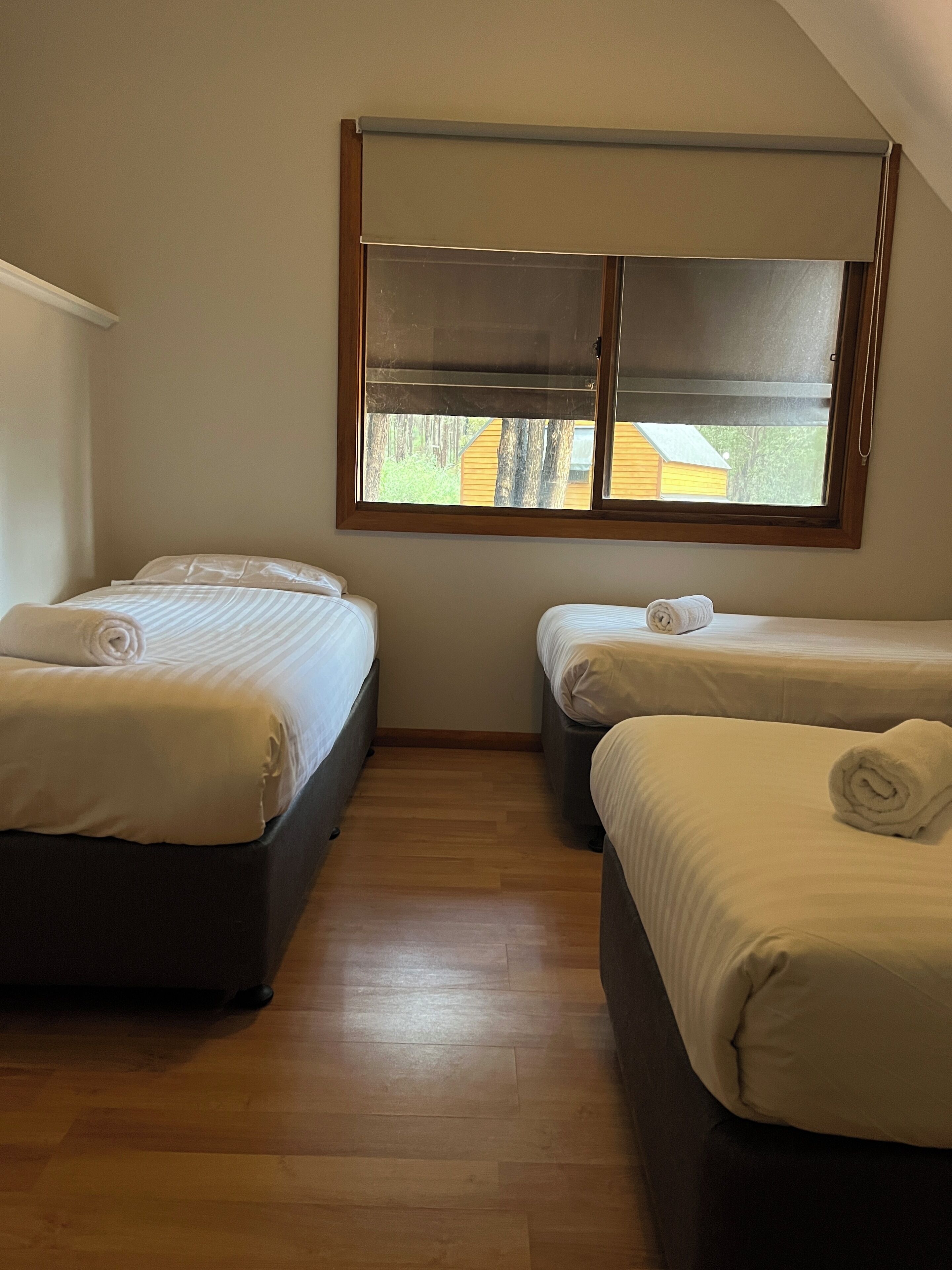 A room with two beds, wooden flooring, a window with blinds, and a view of a building outside.
