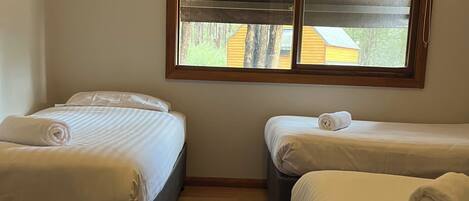 A room with two beds, wooden flooring, a window with blinds, and a view of a building outside.