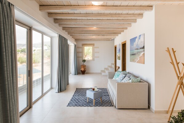 A spacious living room with large windows, a sofa, a small ottoman, and a painting of a beach scene. - Villa Galatia I Free*heated pool, ideal for groups (Kissamos)