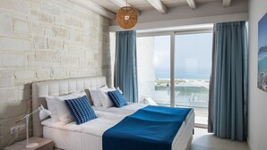 A bedroom with a large bed, a nightstand, a hanging light fixture, and a view of the sea through a window with curtains.