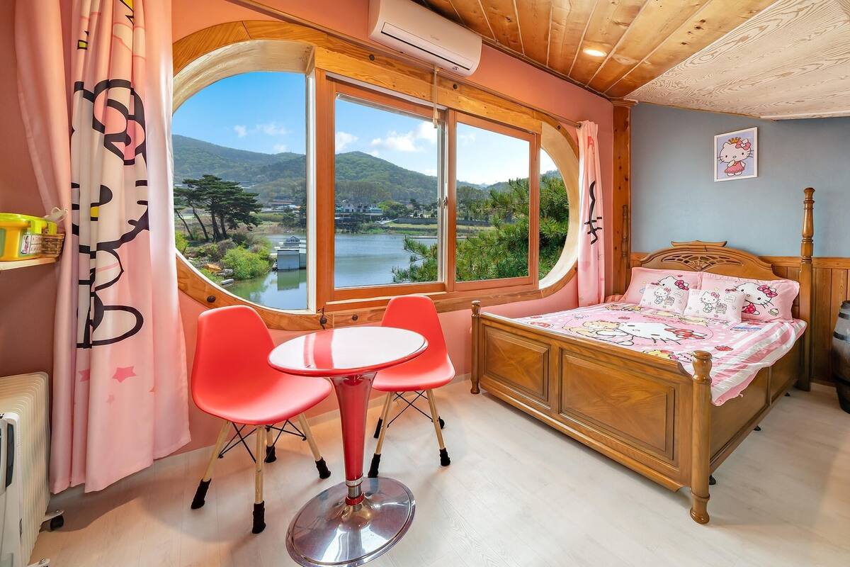 A cozy bedroom with a wooden bed, a round table, and two chairs. A large window offers a scenic view of a lake and mountains.