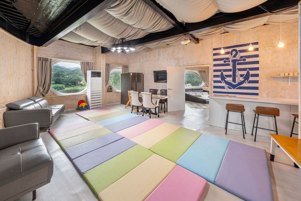 A modern interior with a sofa, colorful floor cushions, a dining area, and a view through a window.