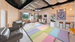 Un interior moderno con un sofá, cojines de colores en el suelo, una zona de comedor y vistas a través de una ventana.