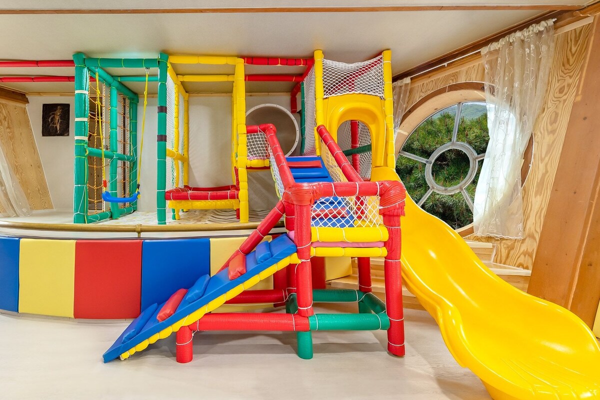 Zona de juegos infantiles con tobogán, red de escalada y un ventanal circular.