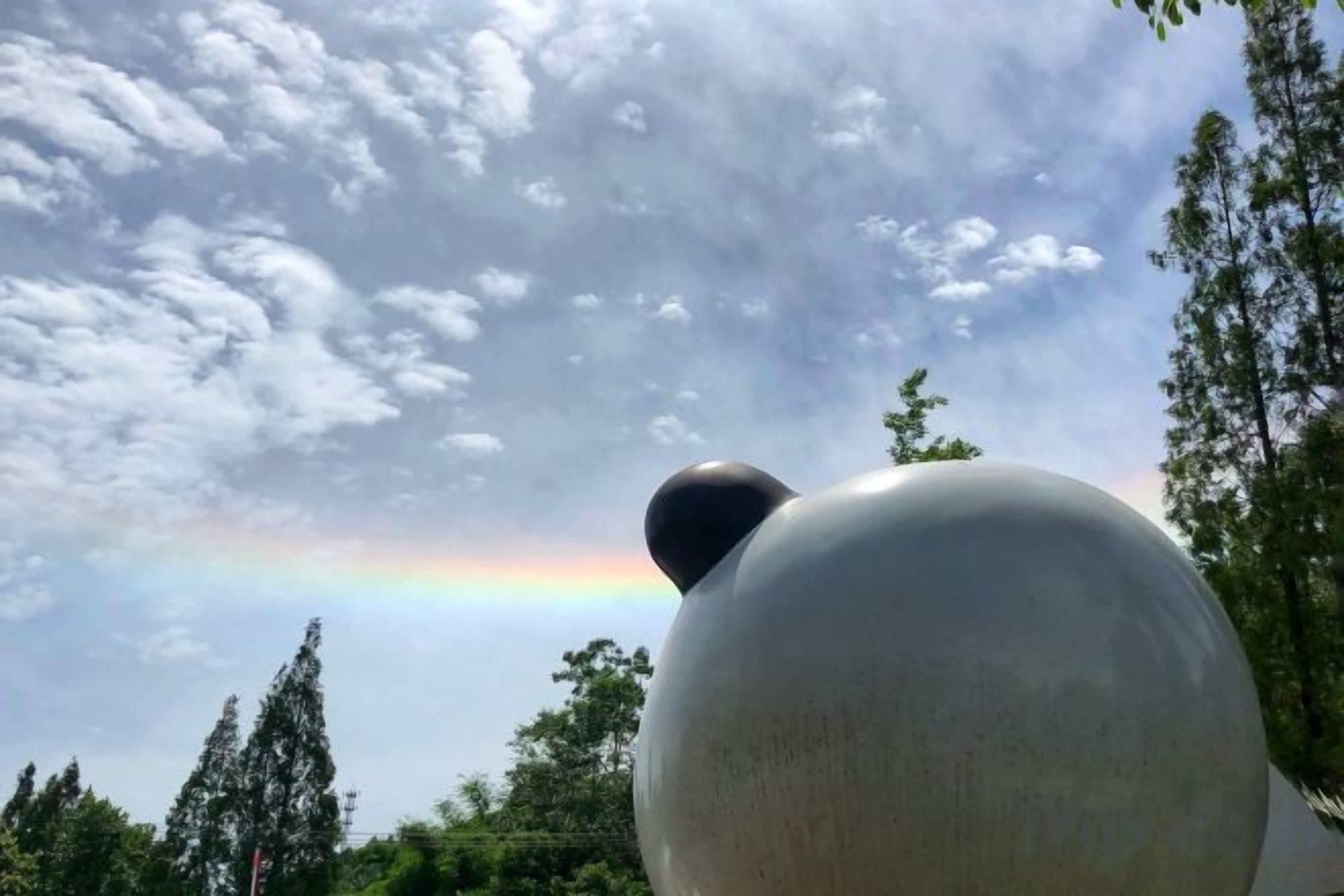 Een grote bolvormige sculptuur met een kleinere bol bovenop, voor een achtergrond van een bewolkte lucht en een regenboog.