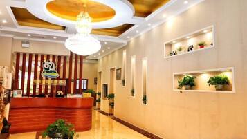 Uma área de recepção de hotel com um balcão de madeira, um candelabro e prateleiras fixadas na parede com plantas.