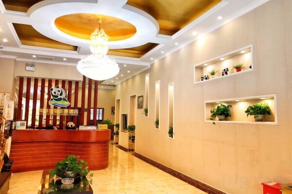 Zona de recepción de un hotel con un mostrador de madera, una lámpara de araña y estantes con plantas.