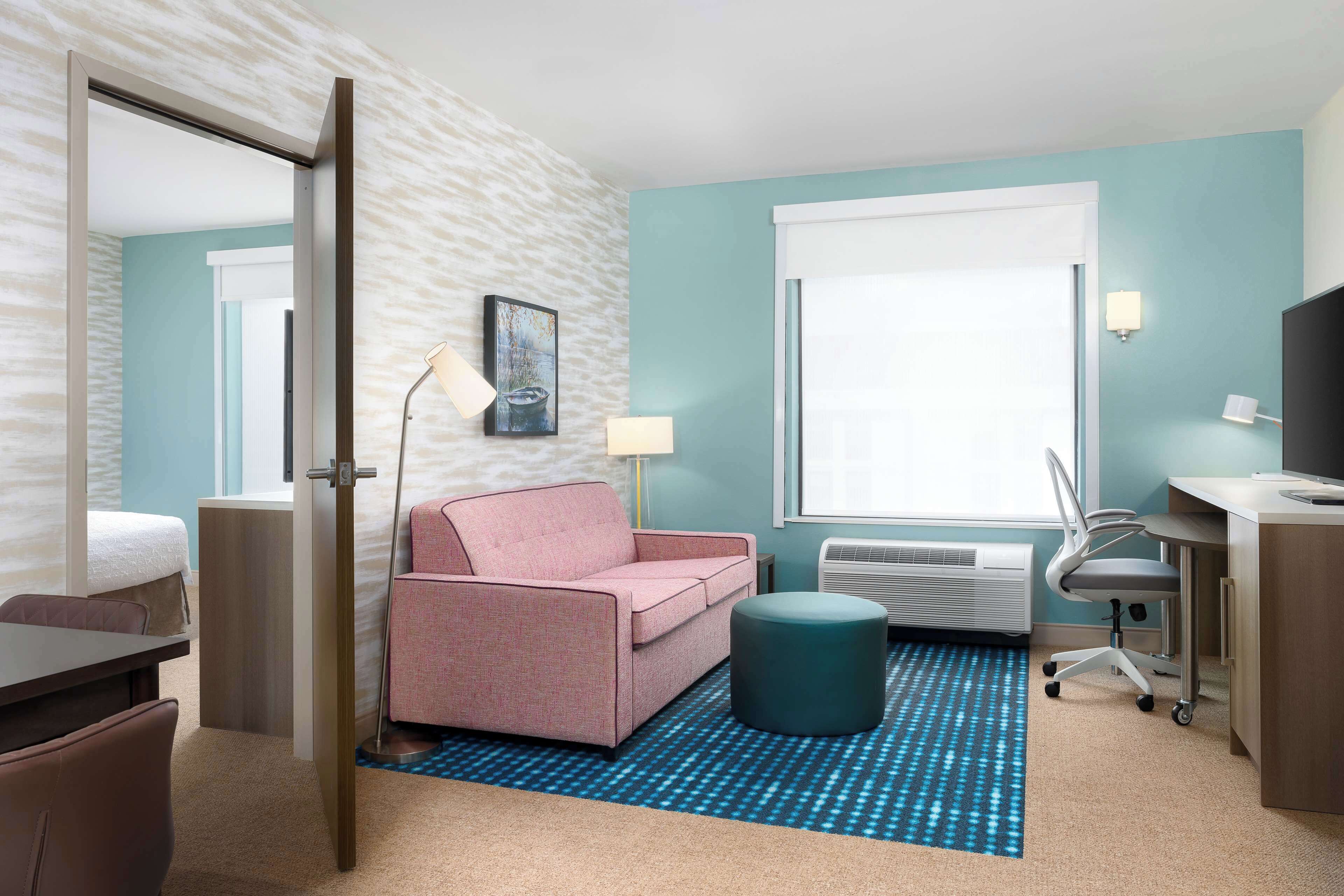 A hotel room with a bed, a pink sofa, a desk with a chair, a standing lamp, and a window with blinds.