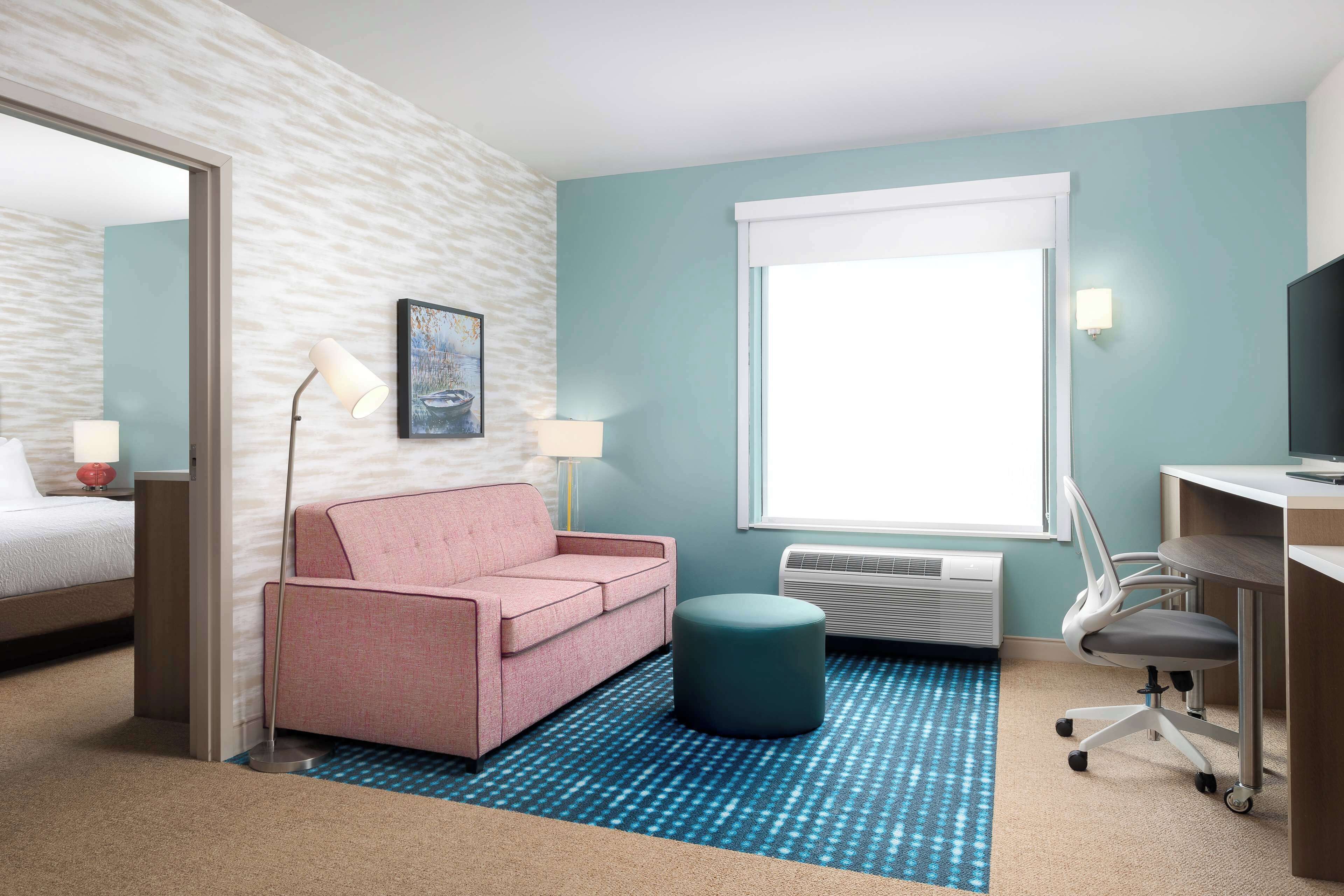 A hotel room with a pink sofa, a desk with a chair, a large window, and a blue patterned rug.