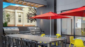 Une terrasse extérieure avec un barbecue, des chaises jaunes et grises, et des parasols rouges.