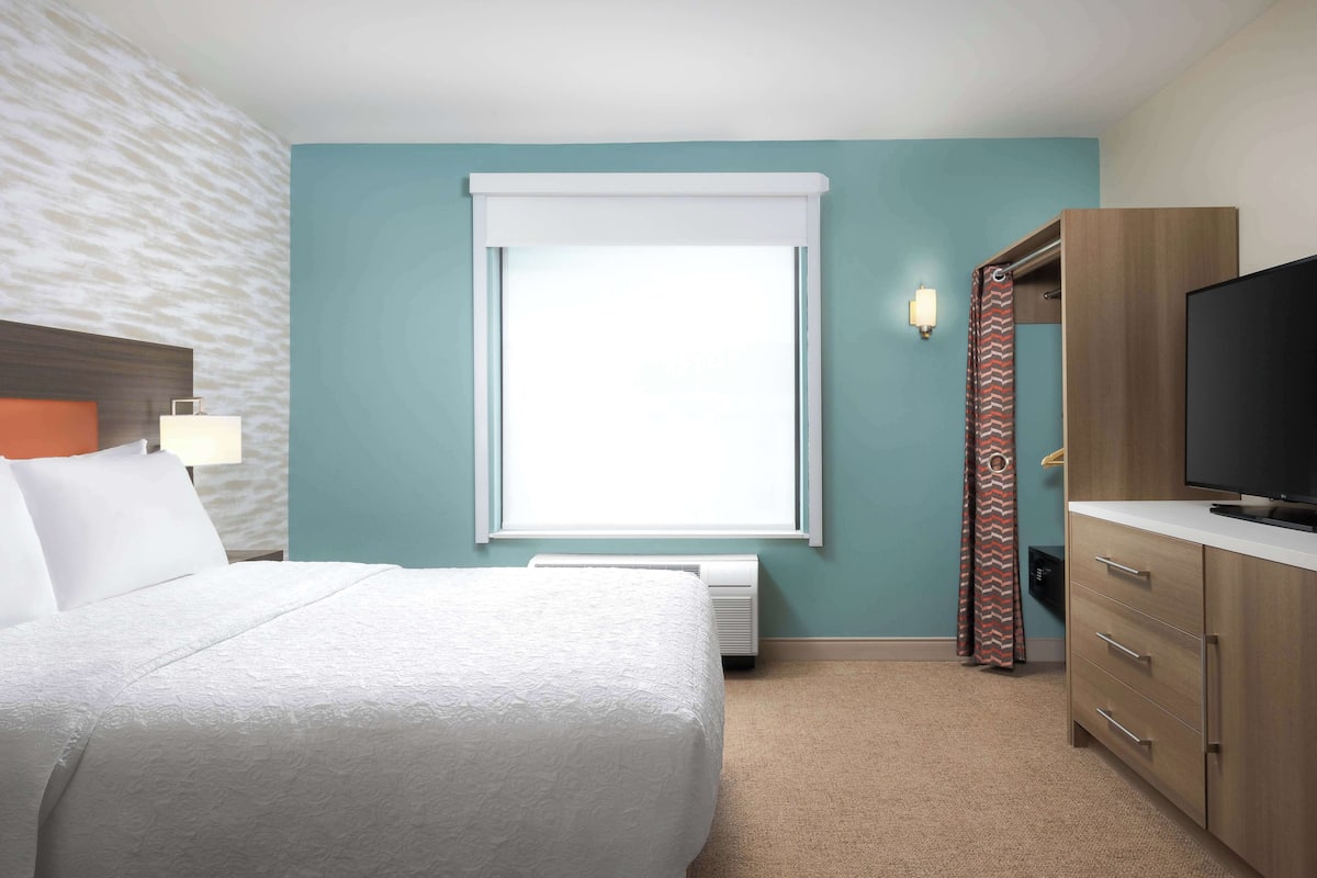 A hotel room with a bed, a television, a window with curtains, and a wall-mounted heater.