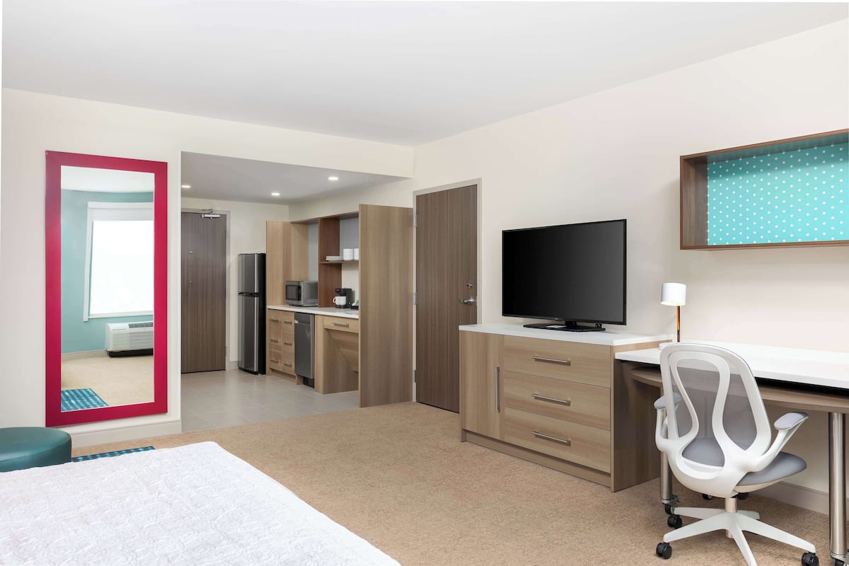 A modern hotel room with a bed, a desk with a computer and chair, a TV, and a kitchenette.