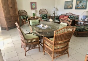 A dining area with a round glass table surrounded by wicker chairs with cushions. There are two side tables with lamps and decorative items.