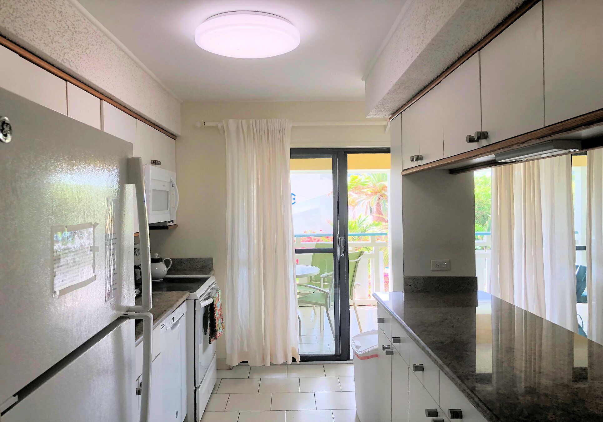 A narrow kitchen with white cabinets, a refrigerator, and a microwave. There is a sliding glass door leading to an outdoor area with a table and chairs.