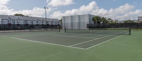 Una cancha de tenis con red, rodeada por una cerca y edificios al fondo.
