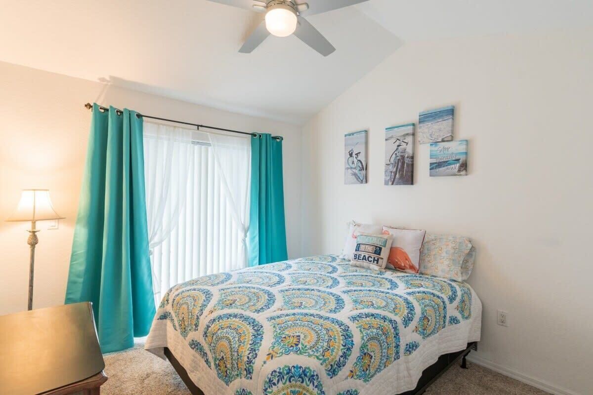 A bedroom with a bed, a ceiling fan, a window with curtains, and wall art.