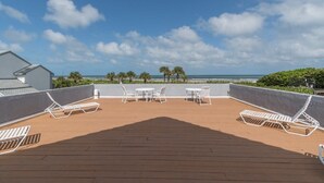 Terraza de madera con sillones y mesas blancos, con vista a una playa con palmeras y el océano.