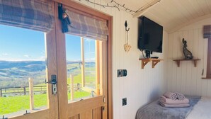 A bedroom with a wooden door, a bed with a grey blanket, a mounted TV, and a view of green fields.