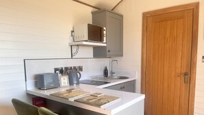 A compact kitchen with a microwave, kettle, and toaster on the counter, a sink, and a wooden door. - Highland View -  a shepherds hut that sleeps 2 guests  in 1 bedroom (Rhayader)