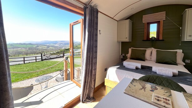 A bedroom with a large sliding glass door leading to a balcony, a bed with multiple pillows, a small table with a decorative item, and a view of the countryside. - Highland View -  a shepherds hut that sleeps 2 guests  in 1 bedroom (Rhayader)