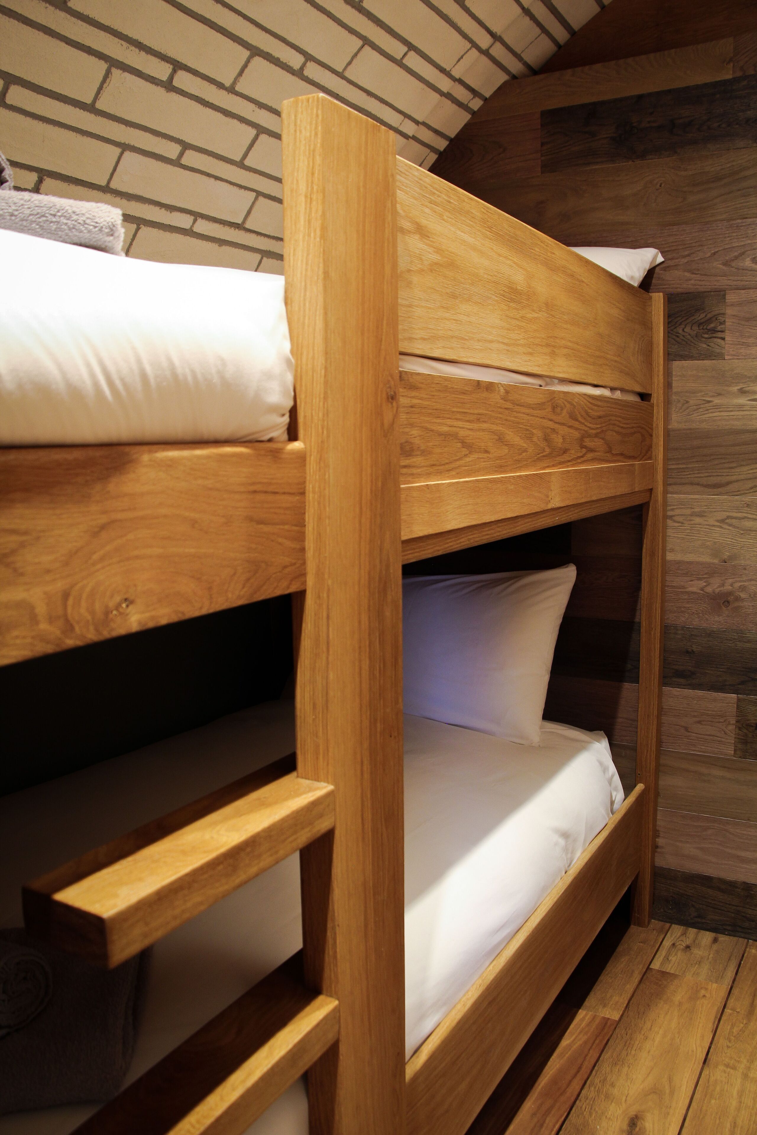 Uma cama de beliche com estruturas de madeira e roupa de cama branca, diante de uma parede de tijolos e soalho de madeira.