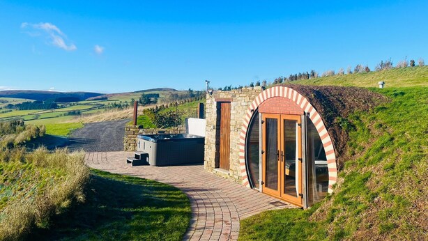 A hot tub and outdoor seating area adjacent to a stone building with a large arched entrance, set in a rural landscape with rolling hills in the background. - Hedgehogs View -  a hobbit hut that sleeps 6 guests  in 3 bedrooms (Rhayader)