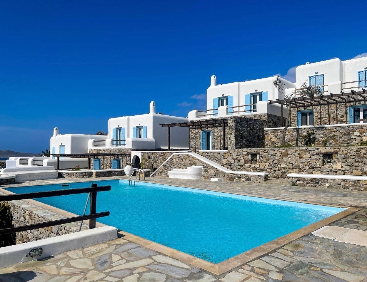 A pool area with white buildings and blue shutters, surrounded by stone steps and a clear blue sky.