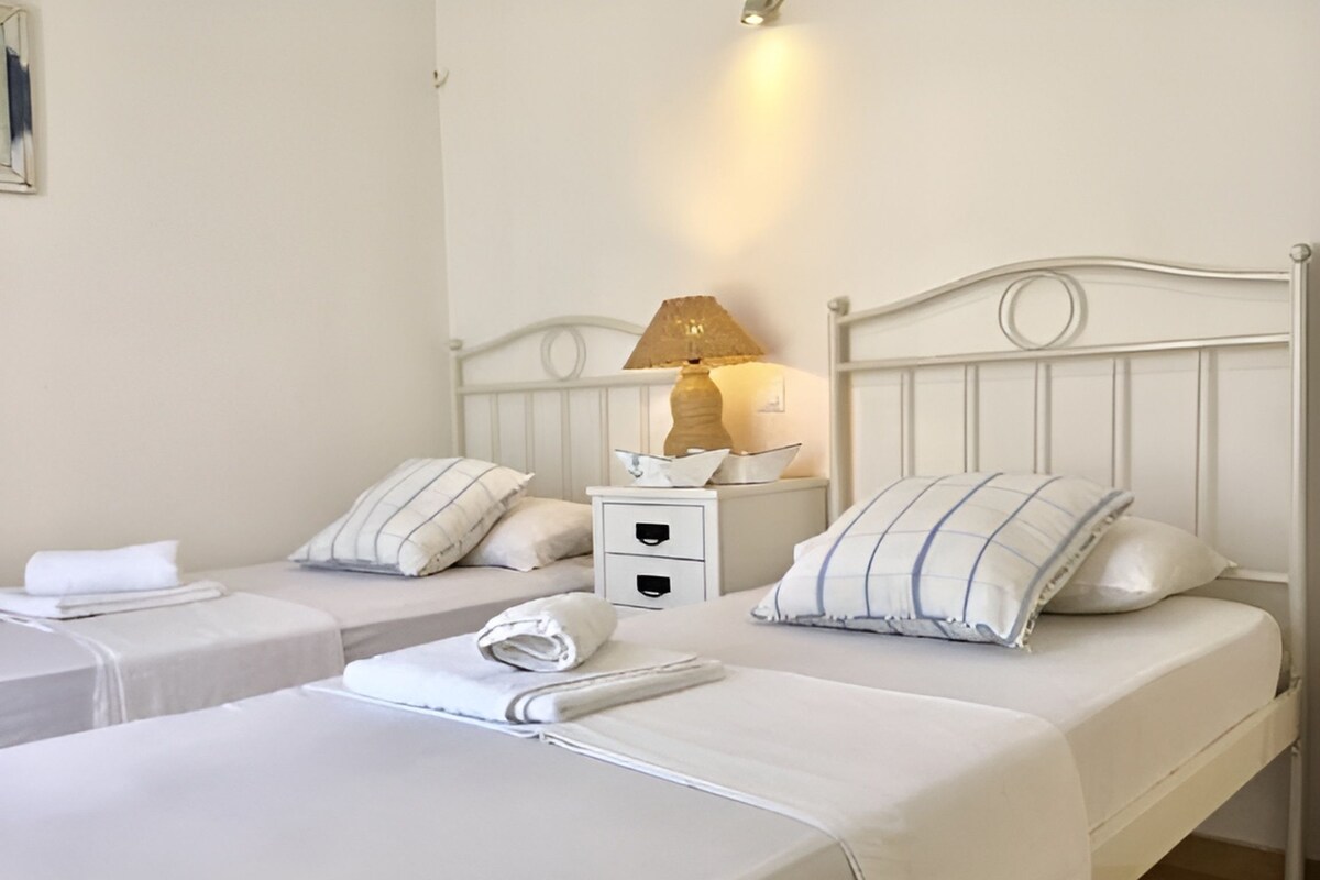 A hotel room with two single beds, each with a pillow and a folded towel. There is a nightstand with a lamp and a small decorative item.