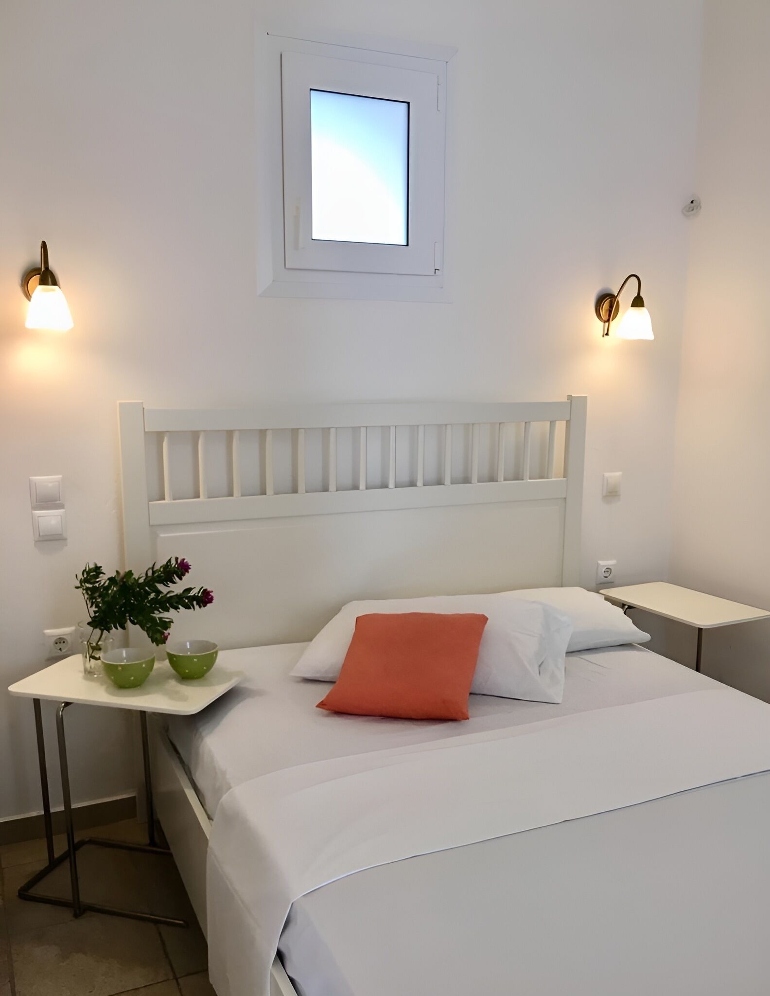 A neatly arranged hotel room with a white bed, a small bedside table with a vase of flowers, and two wall-mounted lamps.