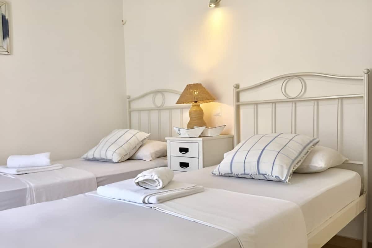Une chambre d’hôtel avec deux lits simples, chacun  étant équipé d’un oreiller et d’une serviette pliée. On y trouve une table de chevet avec une lampe et un petit objet décoratif.