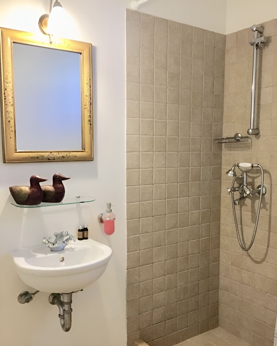 Een badkamer met een wit wastafelmeubel, een spiegel met gouden lijst, een douche met handdouche, en een glazen plank met decoratieve objecten.