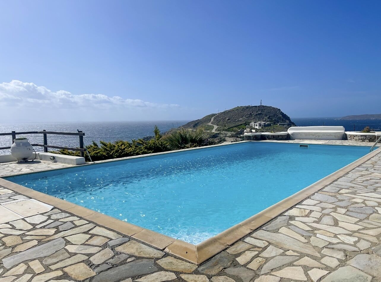 A rectangular swimming pool with a stone-tiled deck, surrounded by greenery and a view of the sea and a hill in the background.