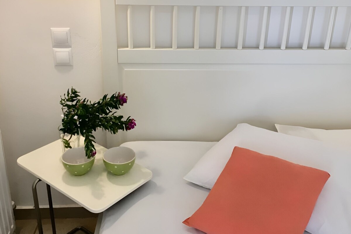 Een net opgemaakt bed met witte en koraal gekleurde kussens, een klein wit nachtkastje met groene schalen en een potplant, en een wandlamp.
