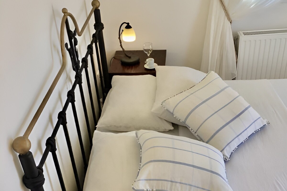 Een keurig opgemaakt bed met witte lakens en gestreepte kussens, een nachtkastje met een lamp, een bord met bananen en een glas water.
