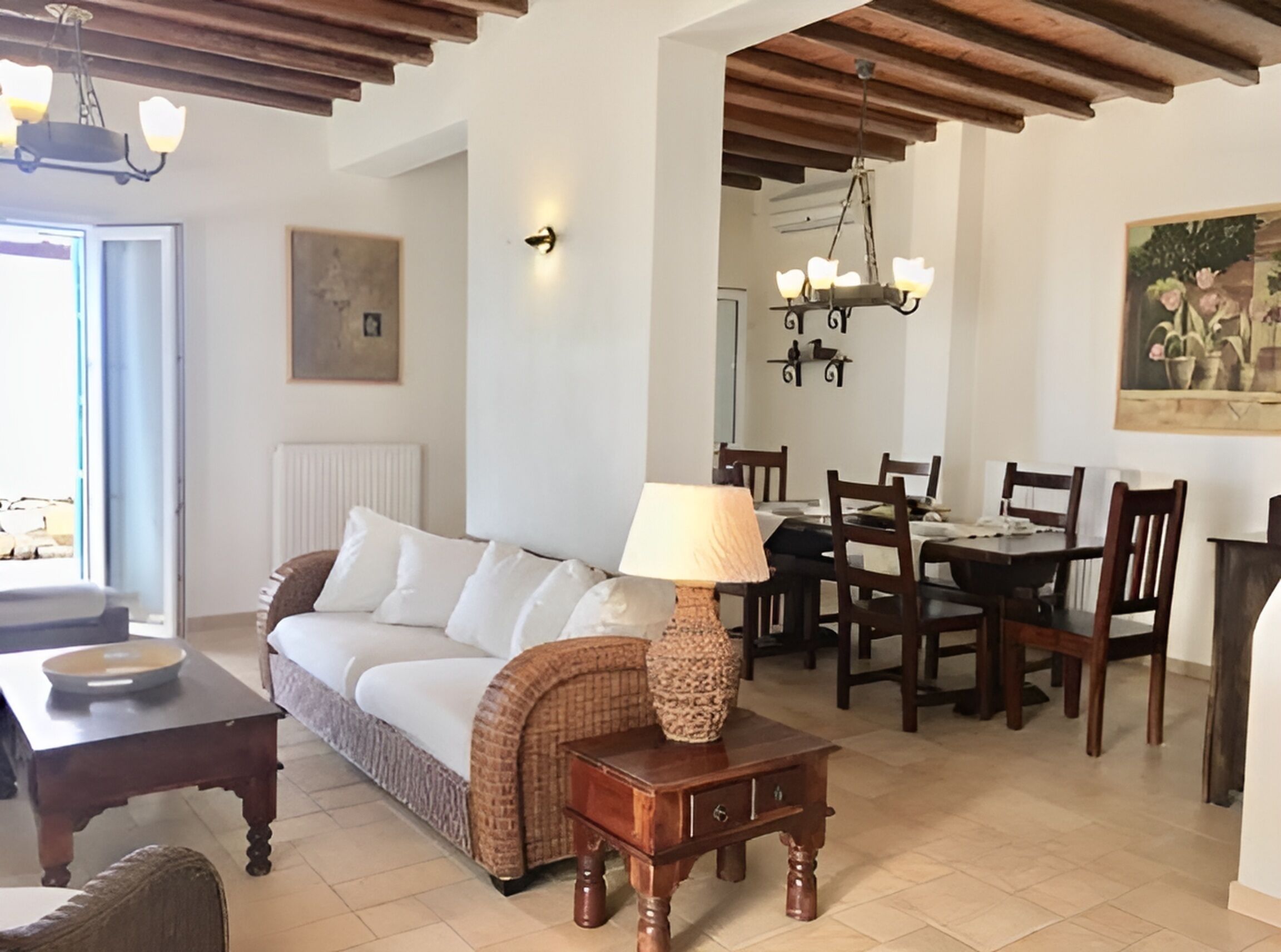 A living room with a sofa, a table with a lamp, a dining area with chairs and a table, and a ceiling with wooden beams.