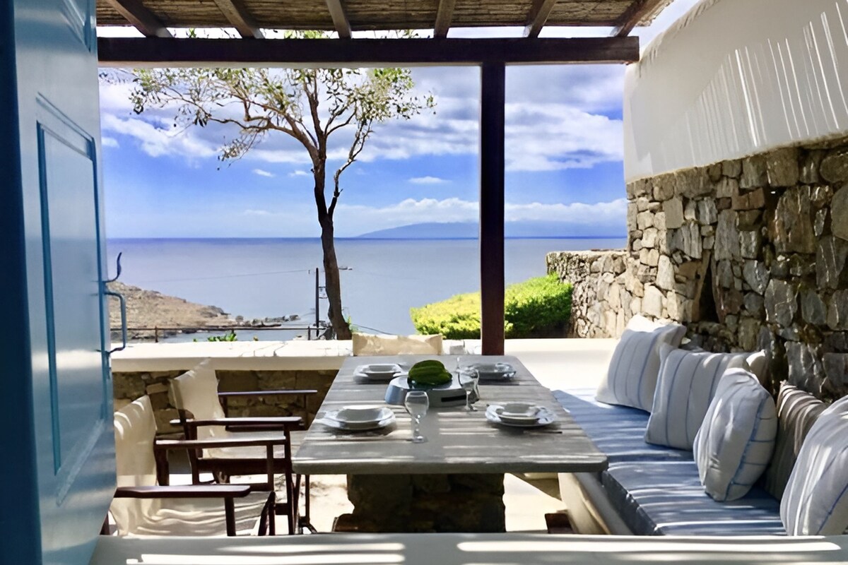 An outdoor dining area with a stone wall, a wooden table set with plates, and a view of the sea.