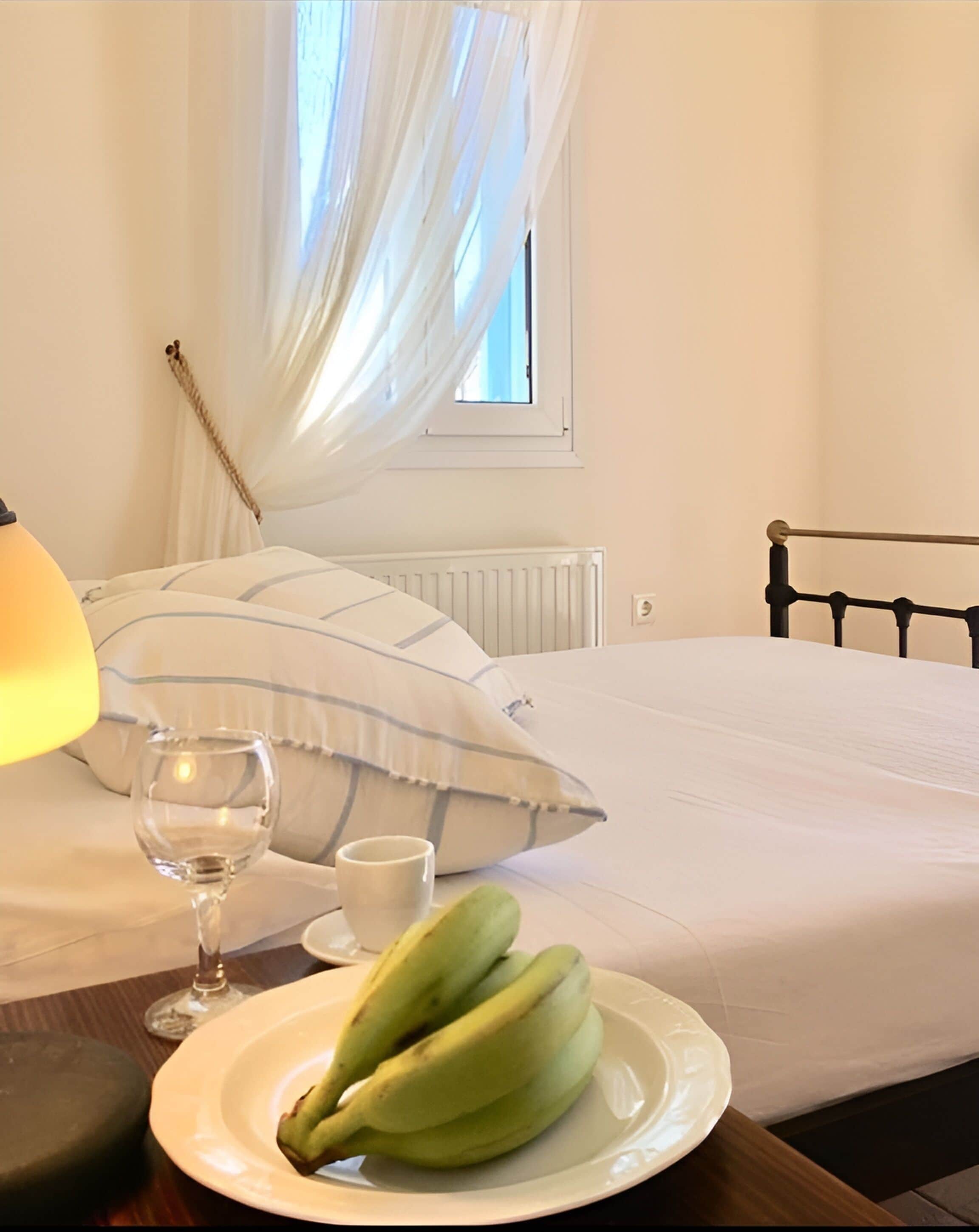 A neatly made bed with white linens, a bedside table with a lamp, a glass of water, a cup, and a plate of bananas.