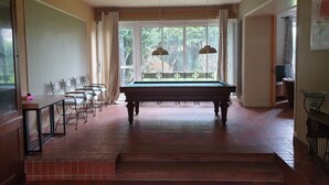 A pool table in a room with a view of greenery outside. There are chairs and a table with a pink object on it.