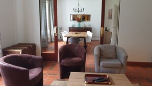 A living room with a wooden coffee table, two armchairs, a dining table with chairs, and a kitchen visible through an open door.