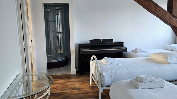 A bedroom with two beds, a piano, a glass table, and a shower.