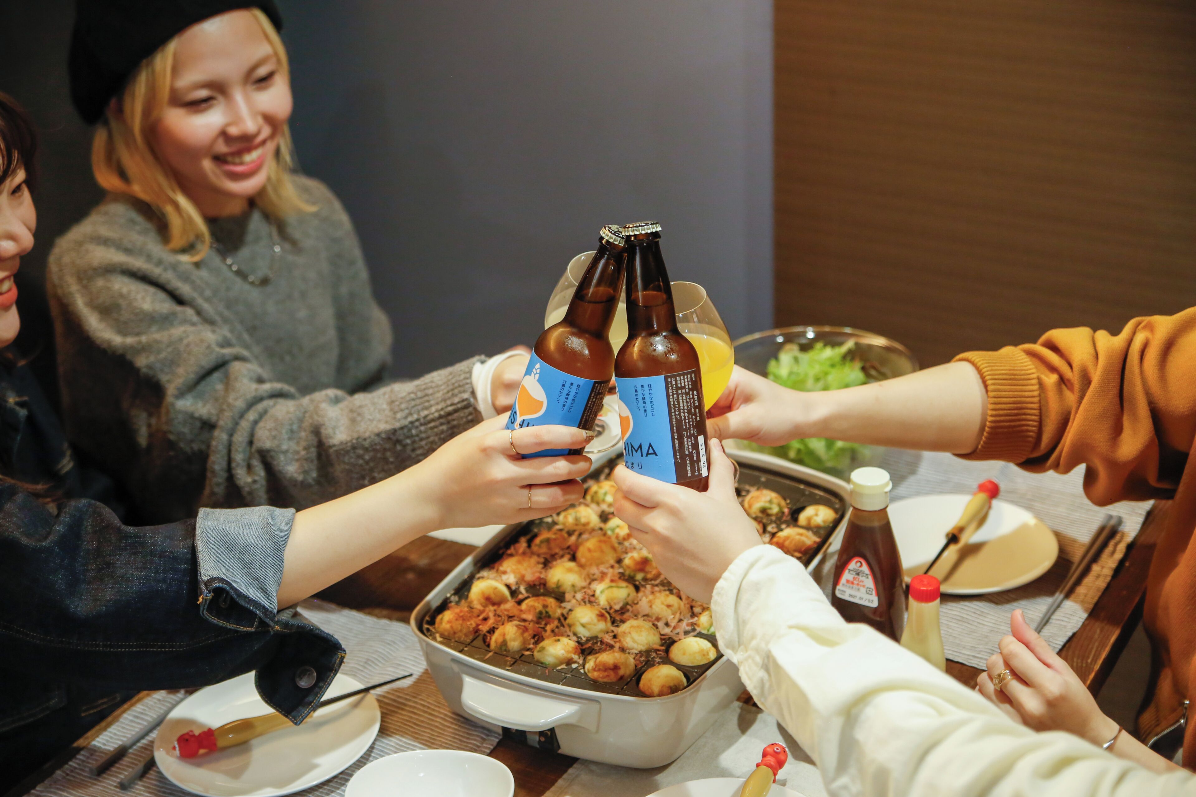 A group of people toasting with beer bottles and glasses, a tray of food, and condiments on a table.