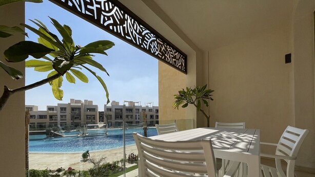 Un balcon donnant sur une piscine et des immeubles. On y trouve une table avec des chaises et un pot de fleurs.