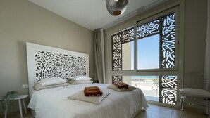 Un lit bien fait, avec du linge de lit blanc et une tête de lit décorative, une petite table de chevet agrémentée d’une plante, et une vue sur l’extérieur à travers des fenêtres à motifs.