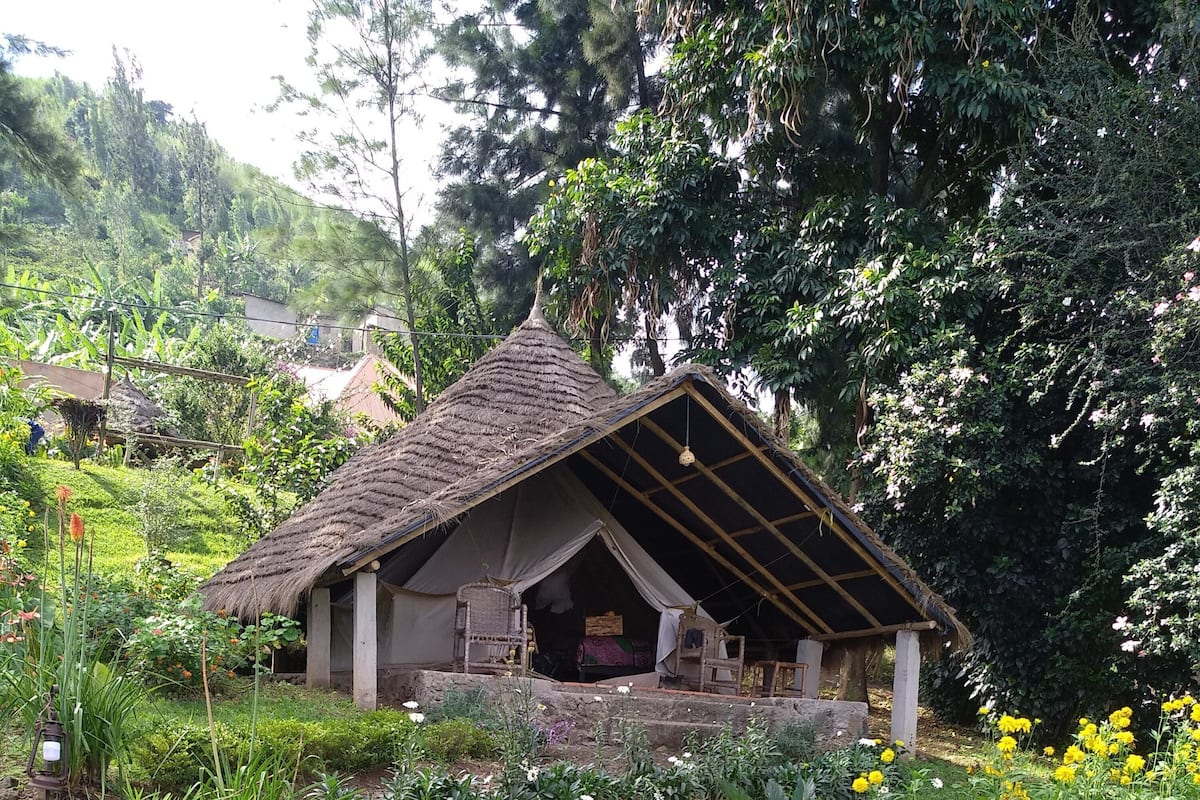 A rustic thatched-roof structure with a simple, open interior, surrounded by lush greenery and flowering plants.