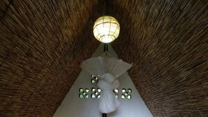 Una habitación con techo de paja, una cama con sábanas blancas, una lámpara colgante y dos máscaras fijadas a la pared.
