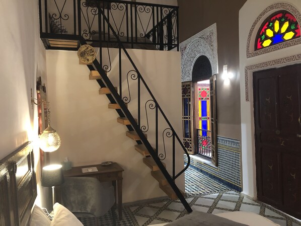 A staircase with ornate black railings, a small table with a lamp, and a view of a room with patterned tiles and a colorful stained glass window.