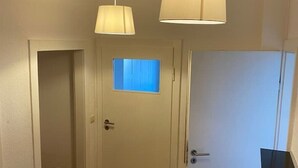 Ein Flur mit Holzboden, weißen Wänden und zwei Türen. Eine Tür hat ein Fenster mit blauen Jalousien, die andere ist geschlossen. Zwei Pendelleuchten sind angebracht.